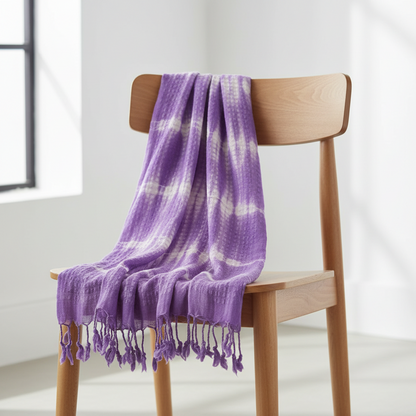 Purple tie-dye blanket with tassels draped over a wooden chair in a bright room.