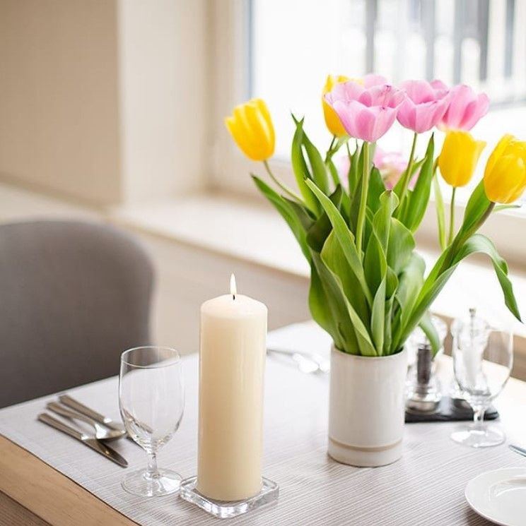 A dining table adorned with a white candle and a vibrant bouquet of yellow and pink tulips, showcasing a Set of 4 Red Pillar Candles (40x80mm - 9 Hour Burn Time) that adds a touch of elegance. Ideal for festive decor and a Christmas gift home fragrance.